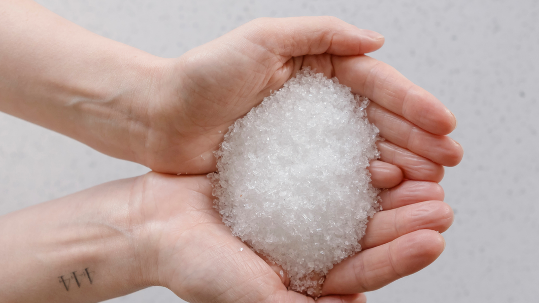Hands holding a scoop of Kunzea Bath Salts crystals over a bath